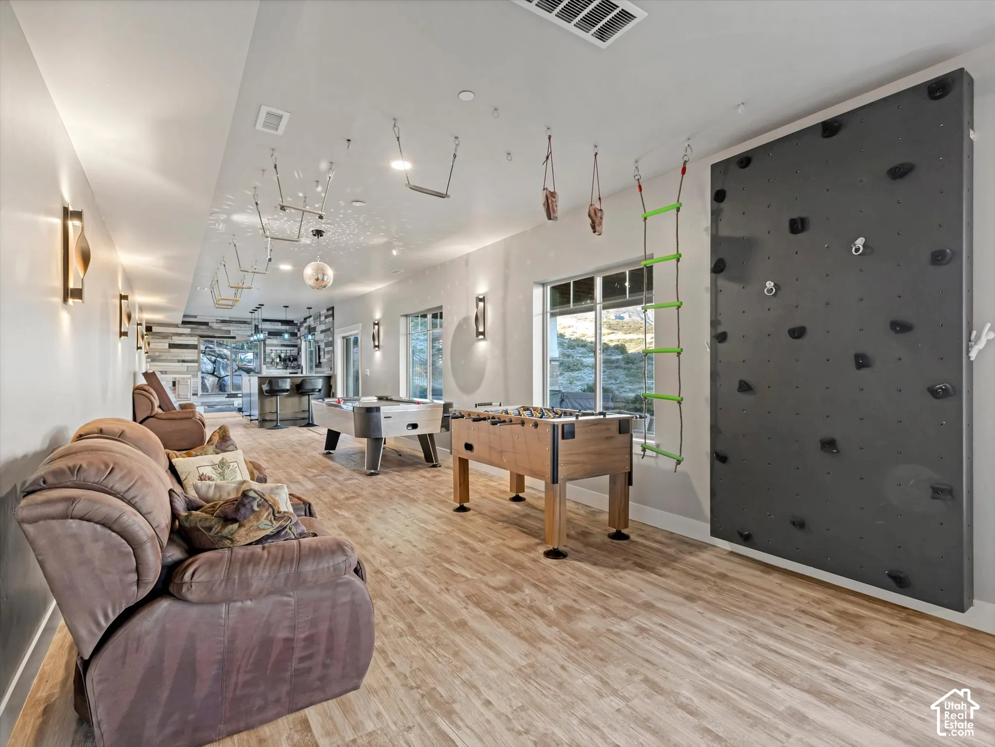 Multi-purpose recreational space featuring wood-finish flooring, a wall-mounted climbing structure, ceiling-mounted exercise apparatus, and recessed wall sconces