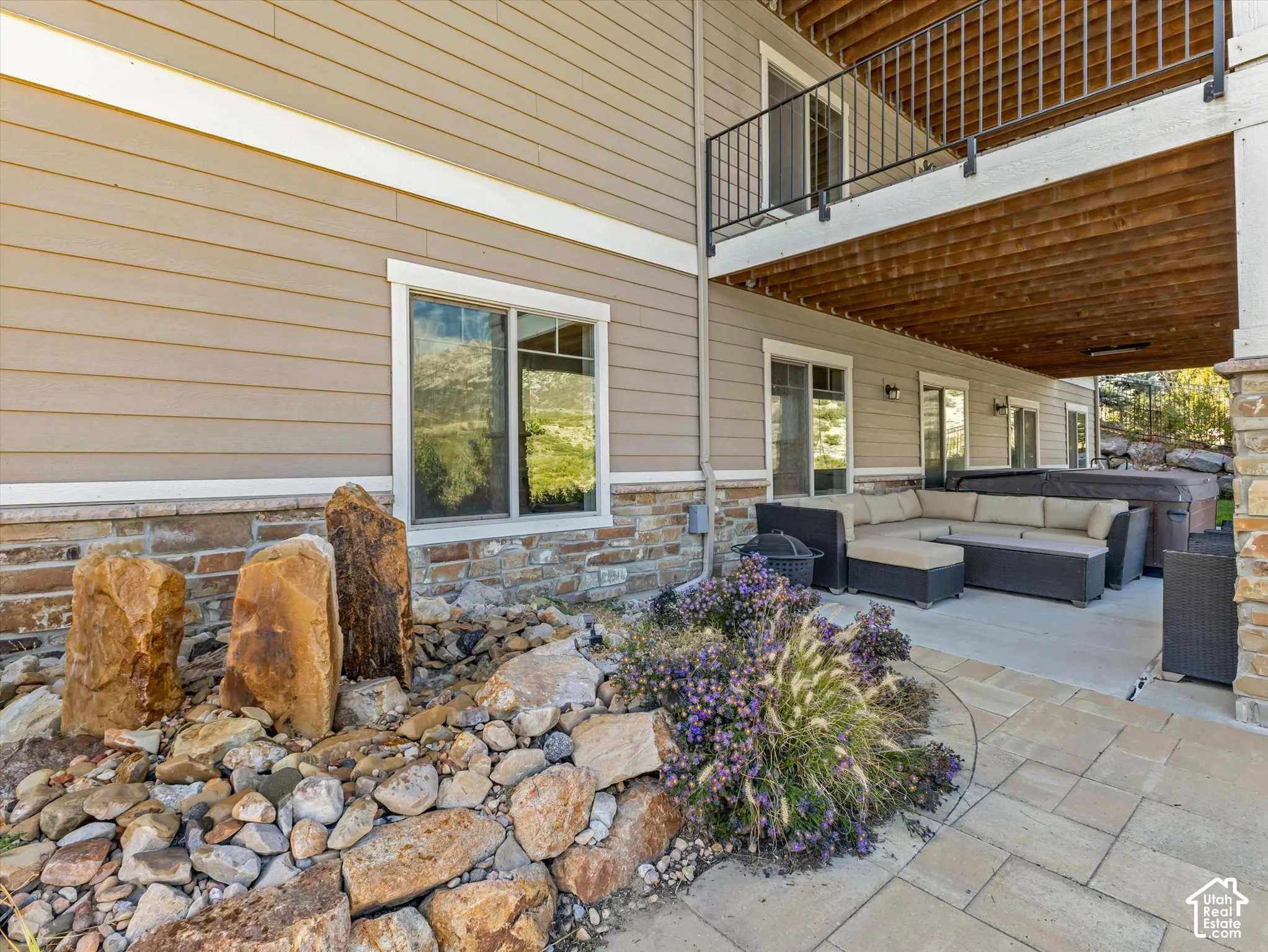 Covered patio with paver flooring, stone veneer foundation, metal balcony railing, and decorative rock landscape feature