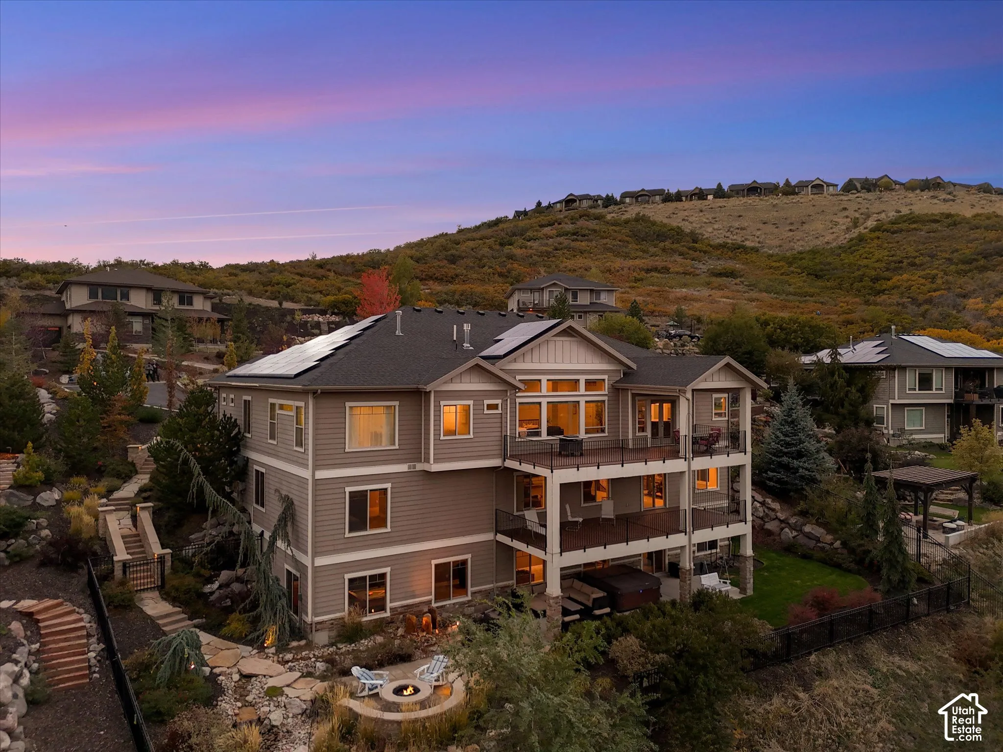 Multi-story residence featuring horizontal siding, rooftop solar panels, stacked-stone accents, tiered wood-finish decks with metal railings, and a stone-lined fire pit area