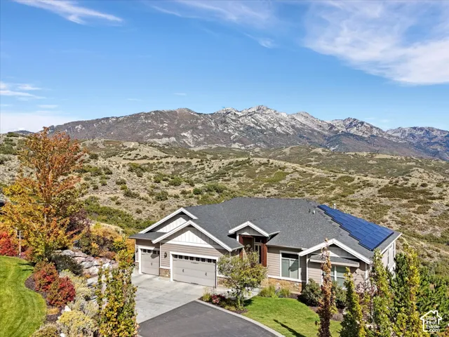 Multi-level residence featuring a multi-car garage, roof-mounted solar panels, stone and horizontal siding exterior, and an asphalt driveway, set against a backdrop of mountain terrain