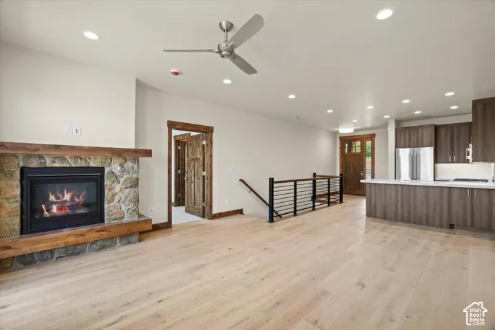 Open-concept living space featuring wood-finish flooring, stone fireplace with a timber mantel, recessed lighting, and a kitchen equipped with dark-finish cabinetry