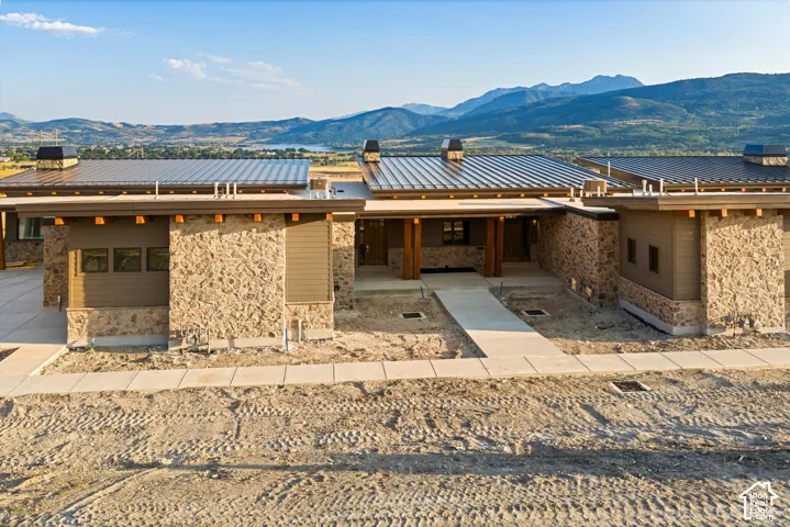 Modern single-story exterior featuring a standing-seam metal roof, stone veneer siding, exposed timber beams, and mountain scenery