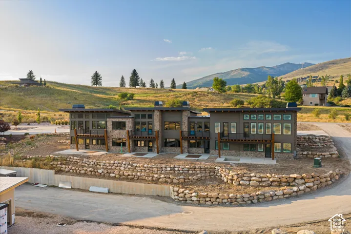 Contemporary multi-unit exterior featuring stacked stone masonry, expansive window arrays, metal railings, and tiered natural rock retaining walls set against a mountain backdrop