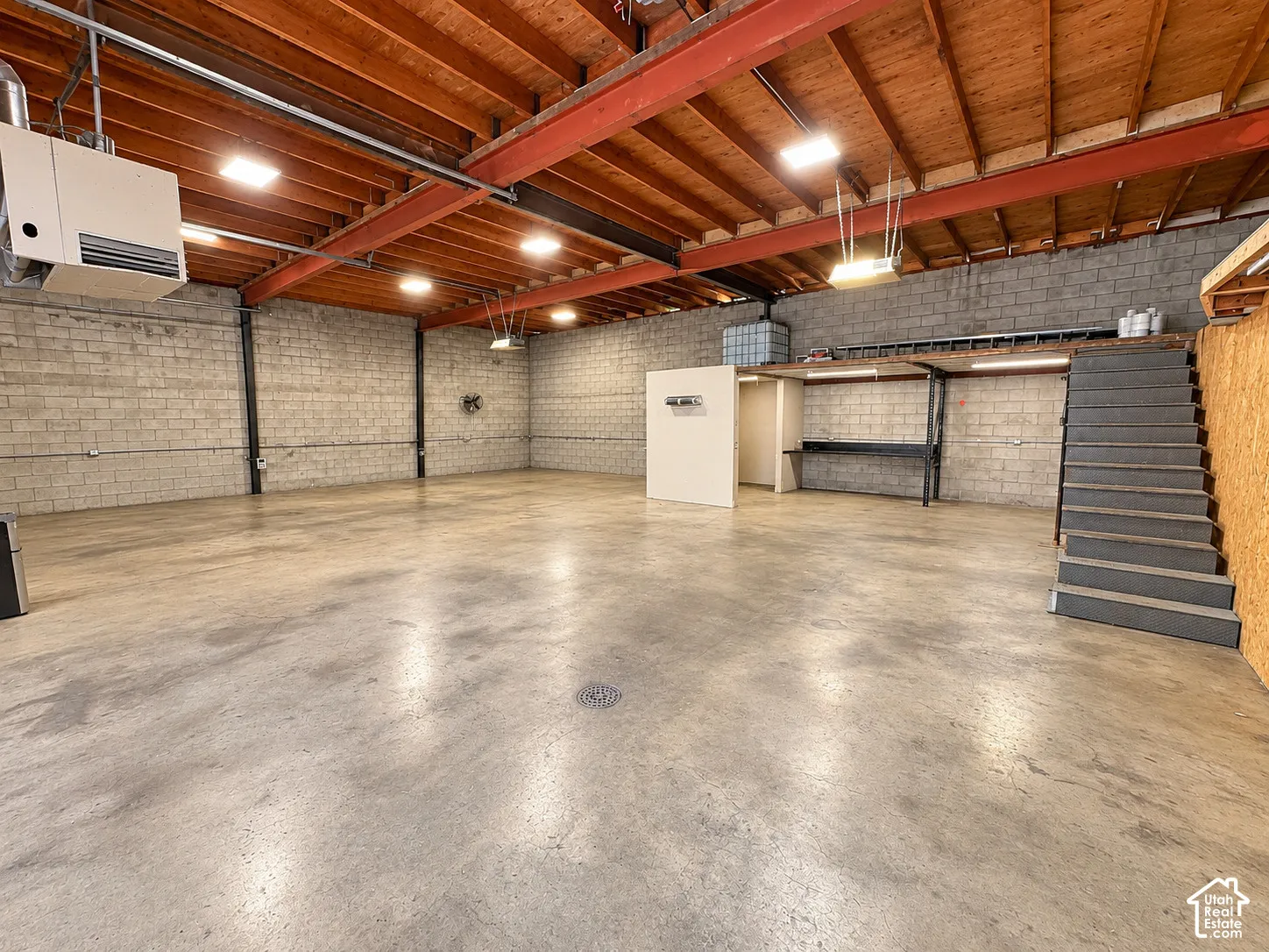 Open-concept industrial space featuring exposed wood-beam ceilings, concrete block walls, durable concrete flooring, and a built-in staircase to a mezzanine level