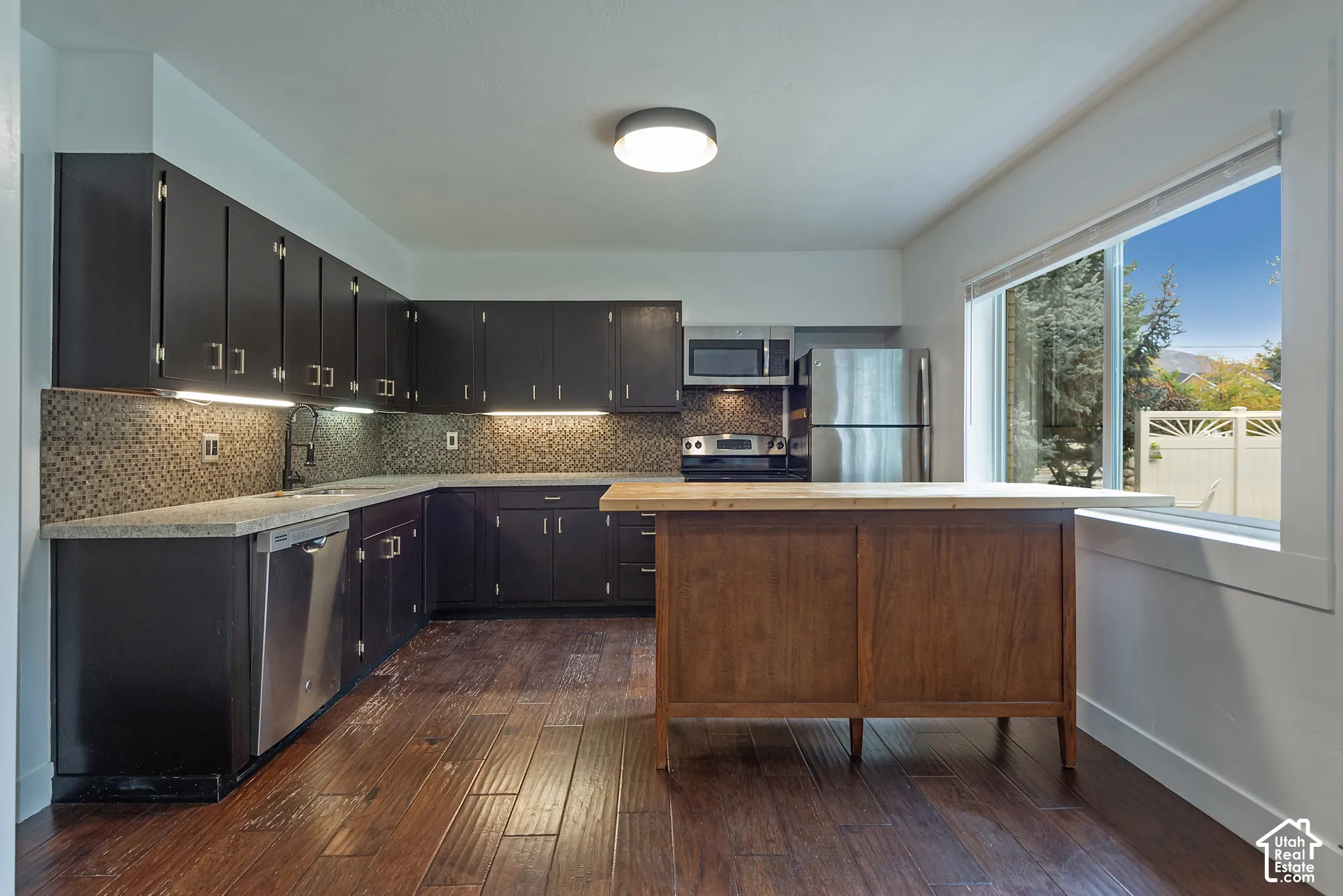 Kitchen featuring dark cabinetry, stone-finish countertops, tile backsplash, wood-finish flooring, and stainless steel appliances