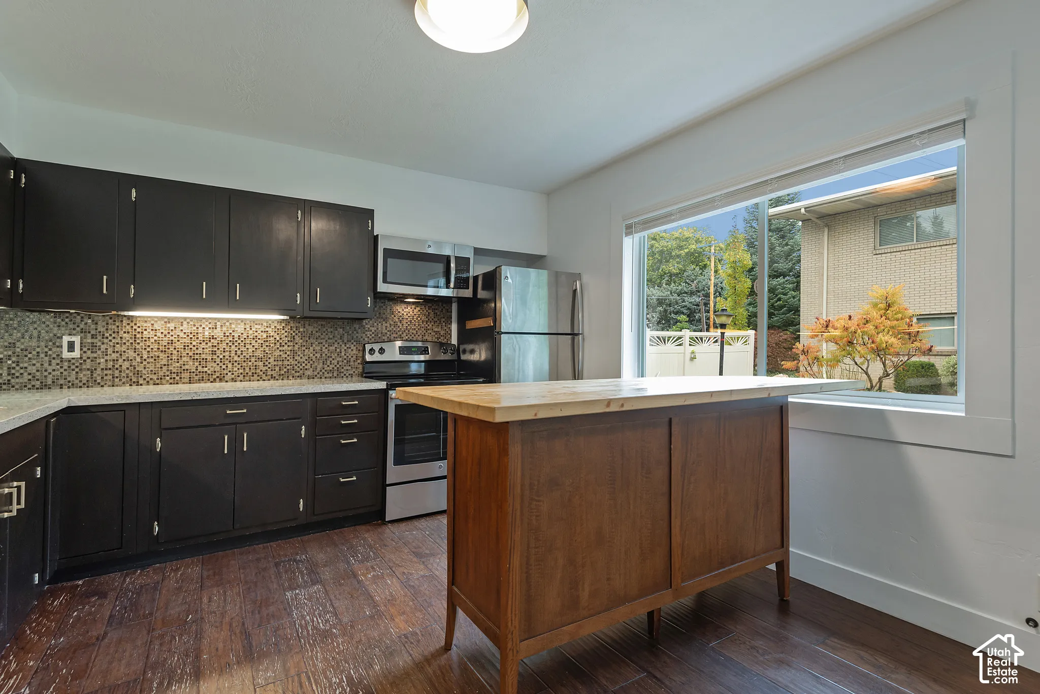 Kitchen featuring dark-finish cabinetry, stainless steel appliances, mosaic tile backsplash, wood-finish flooring, and a central island with a wood-tone surface