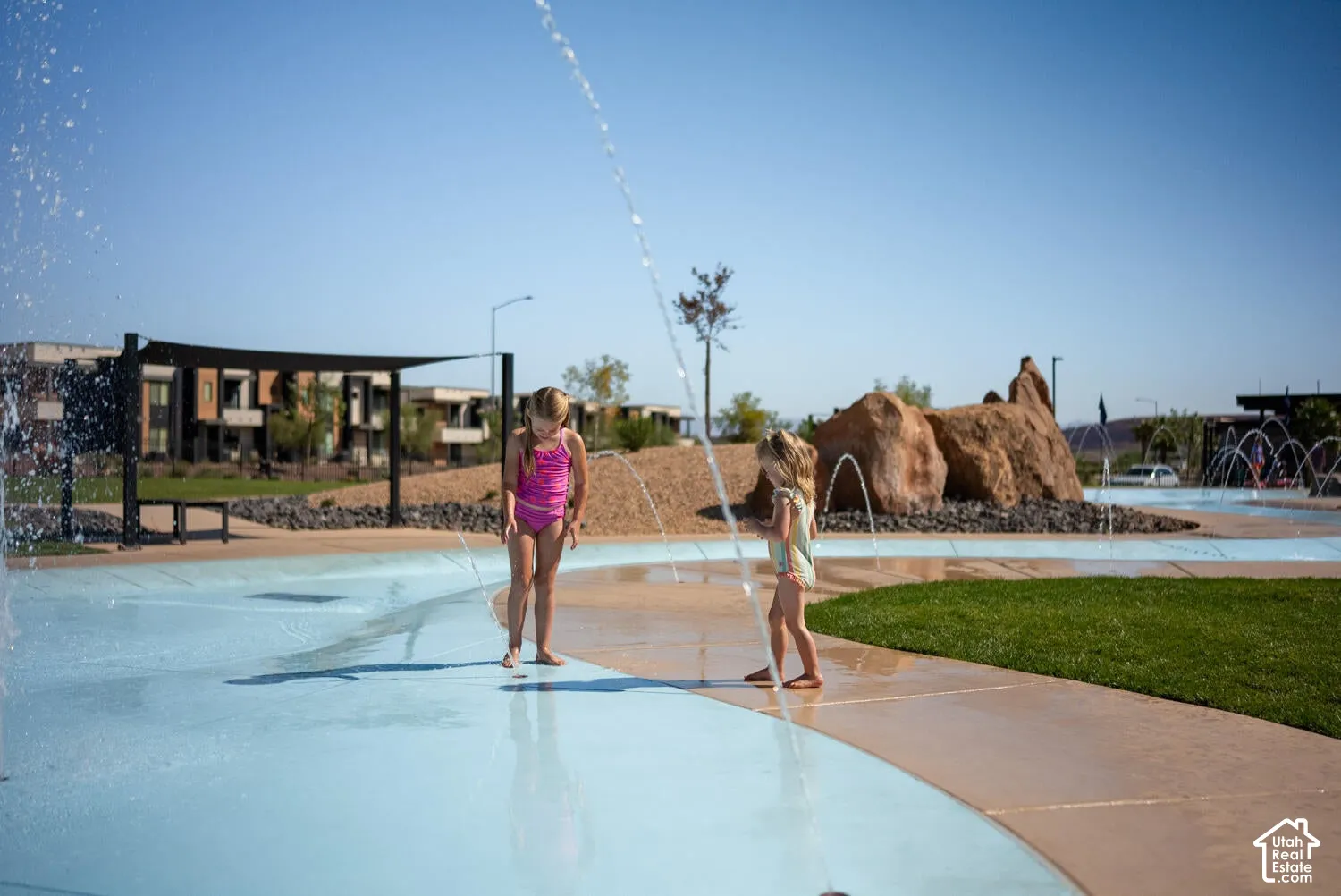 Community splash pad featuring integrated water jets, a concrete basin, manicured lawn areas, and decorative boulder hardscaping
