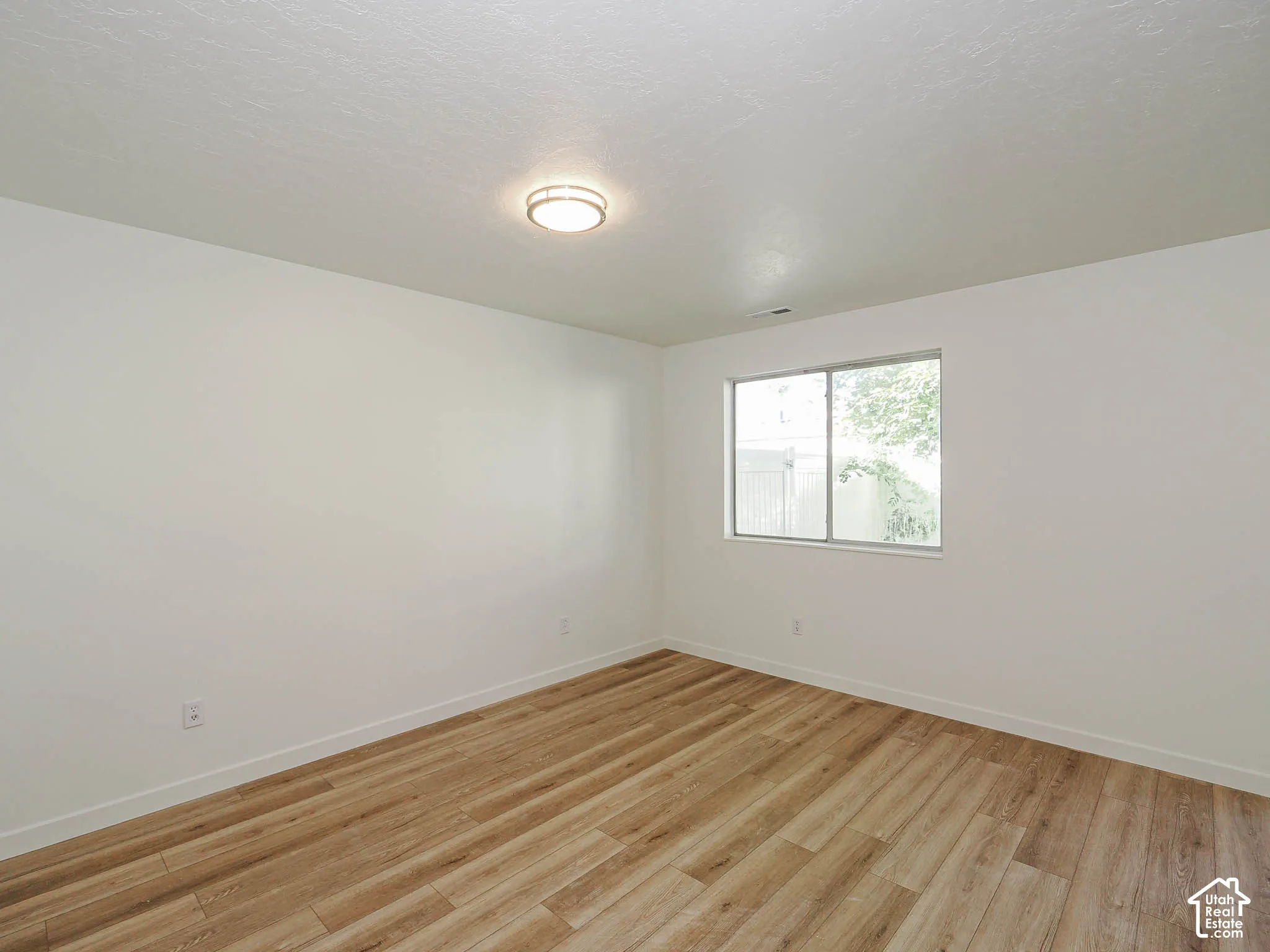 Neutral interior space featuring wood-finish flooring, smooth-textured walls, a single-pane sliding window, and a flush-mount circular ceiling fixture