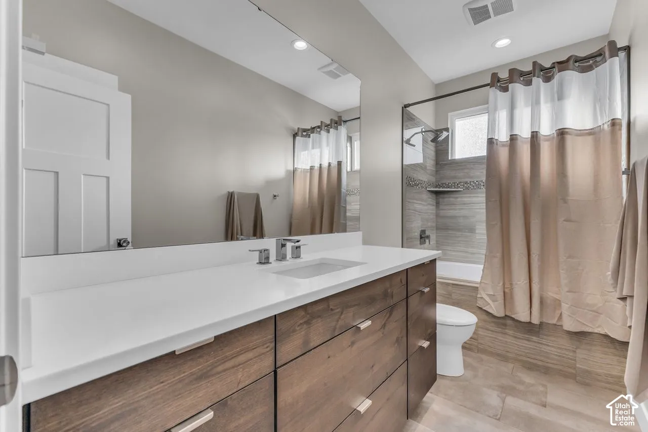 Bathroom featuring a wood-finish vanity with a stone-surface countertop, an undermount rectangular sink, a large wall-to-wall mirror, and a tiled shower surround with built-in shelving