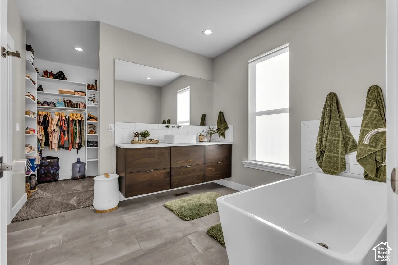 Floating wood-finish vanity with a rectangular vessel sink and white countertop, full-width vanity mirror, freestanding soaking tub, and walk-in closet with built-in shelving