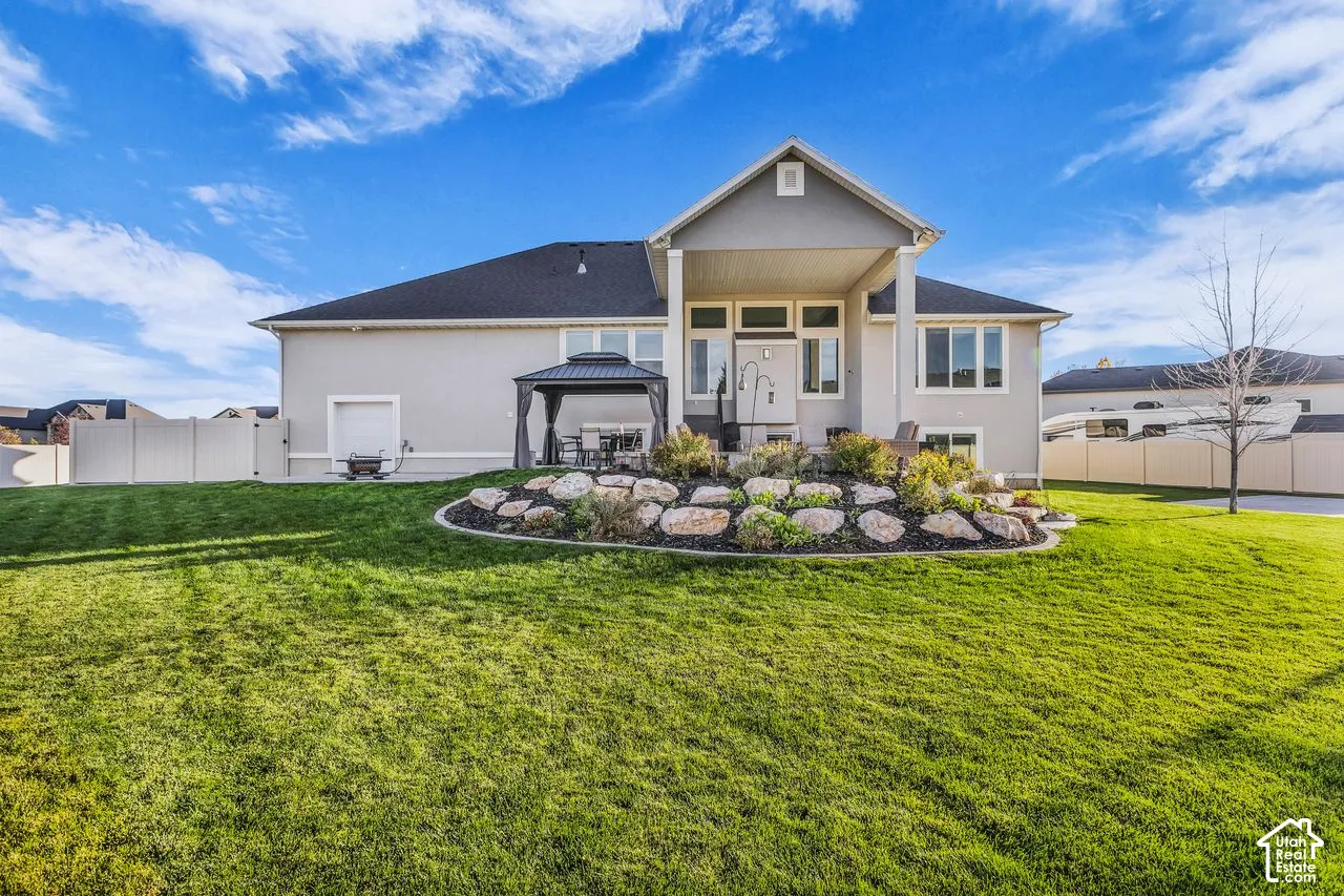 Rear exterior featuring a covered patio, tiered stone landscaping, extensive lawn, and white vinyl privacy fencing