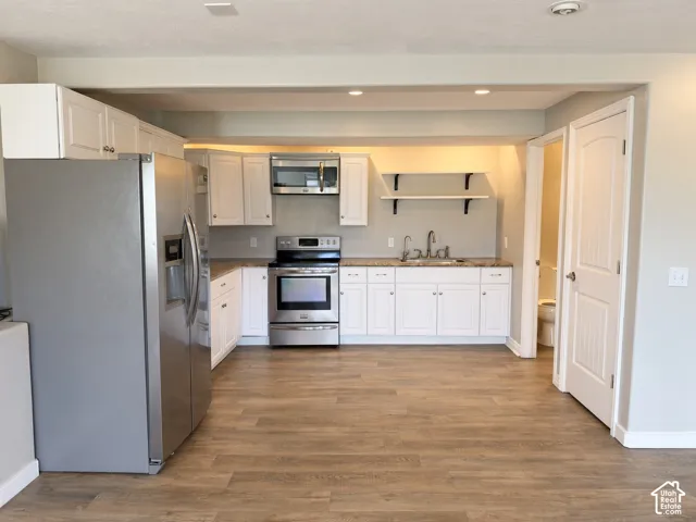 Kitchen featuring white shaker-style cabinetry, stainless steel appliances, wood-finish flooring, floating shelves, and recessed lighting