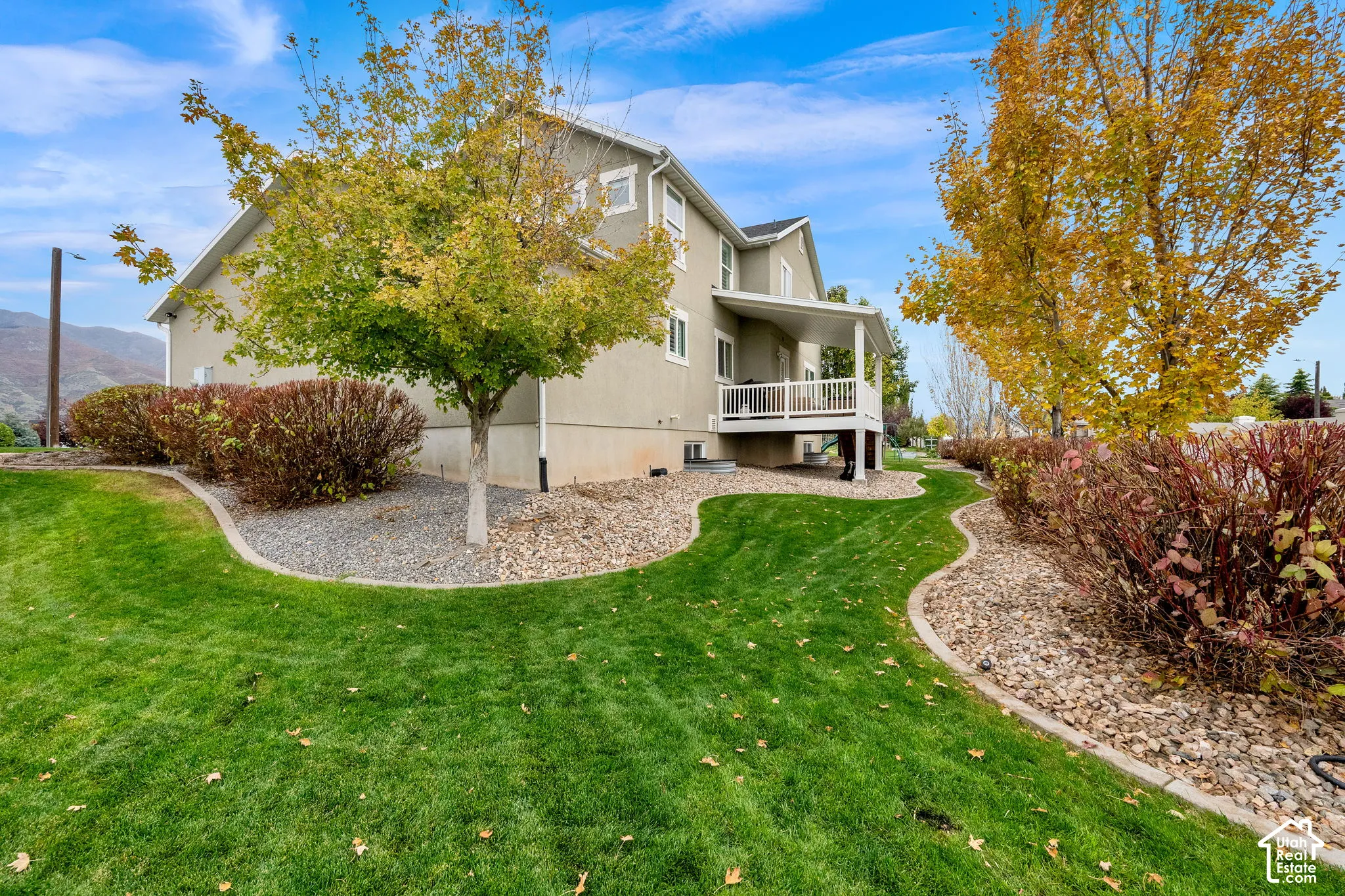 Exterior featuring a covered deck, manicured lawn with curved concrete landscape curbing, decorative rock beds, and established landscaping against a mountain backdrop