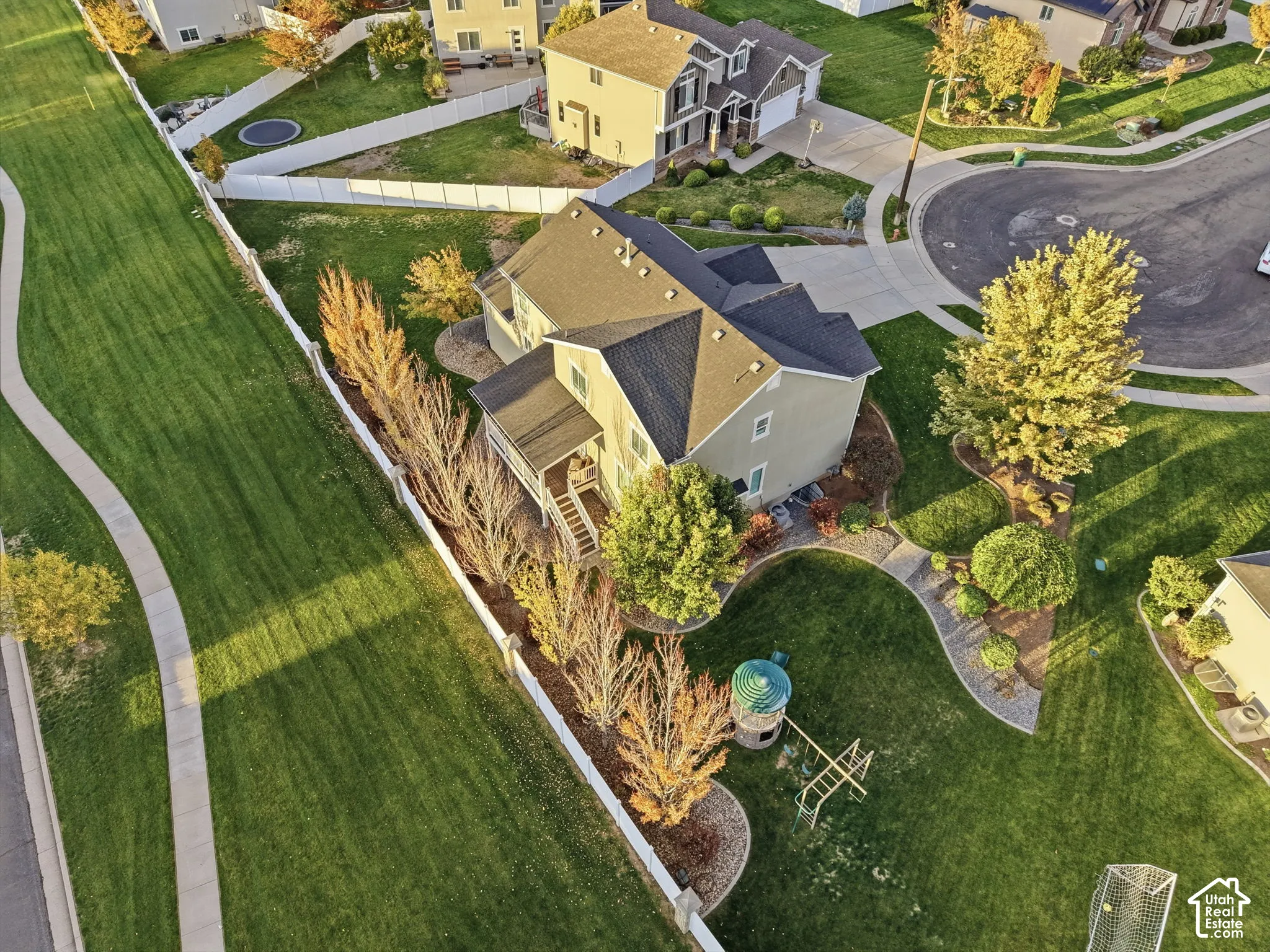 Aerial view of a two-story residence featuring a shingled roof, attached garage, rear wooden deck, and landscaped yard enclosed by white fencing