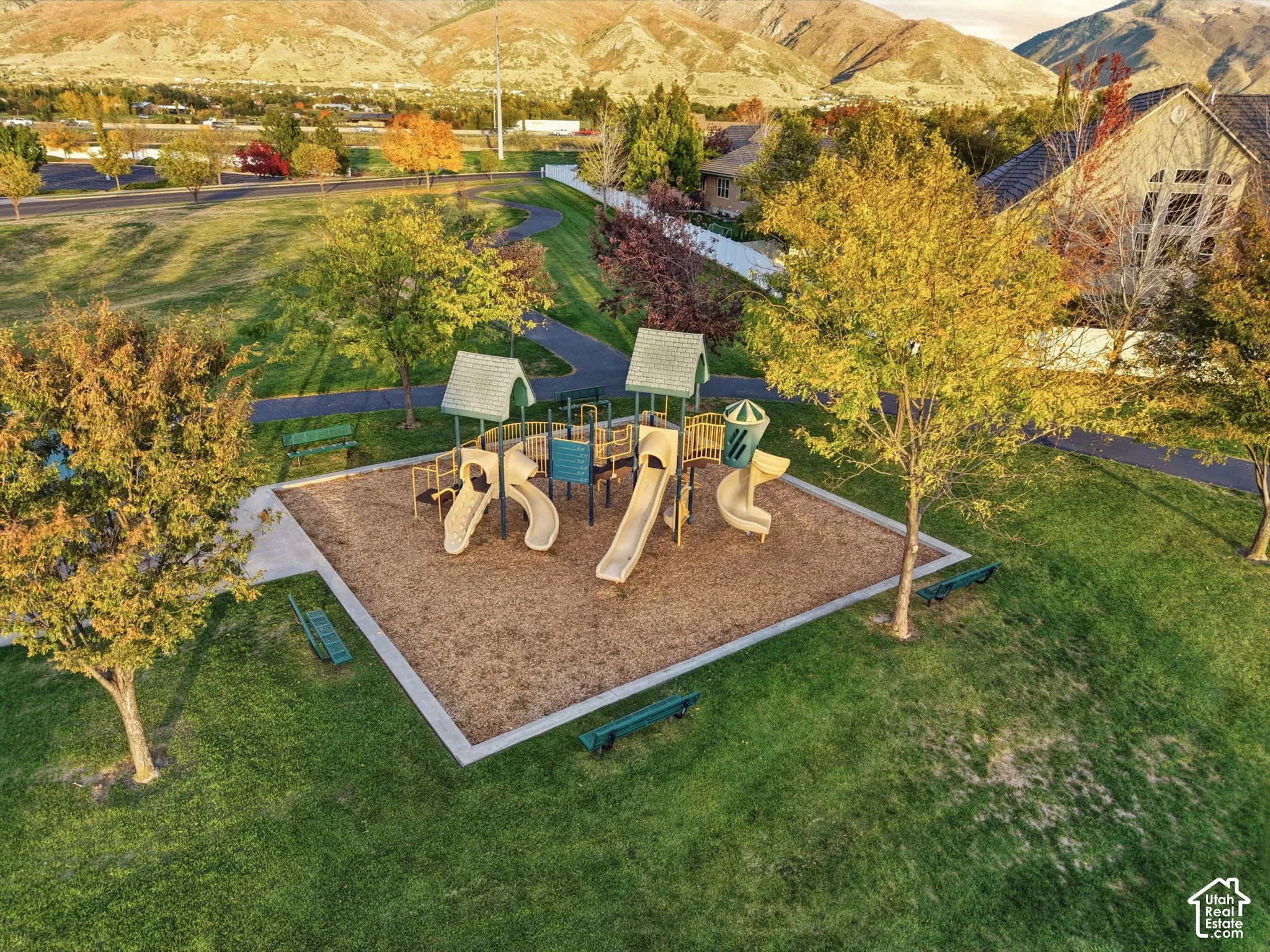 Community park featuring a multi-component playground structure, manicured green space, paved pathways, and mature trees against a mountain backdrop
