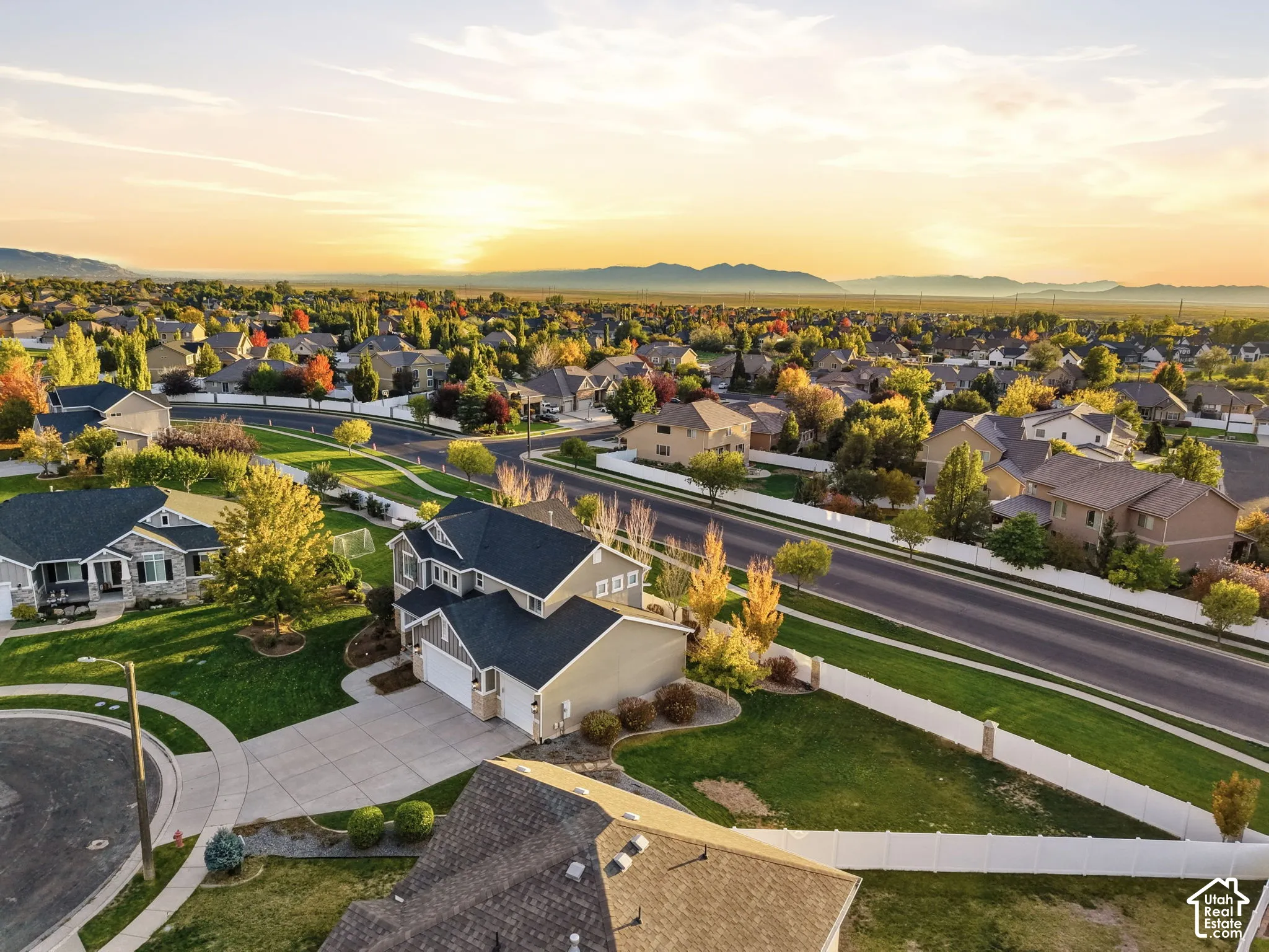 Aerial perspective of a multi-story residence with a multi-car garage, concrete driveway, and expansive lawn area enclosed by white vinyl fencing