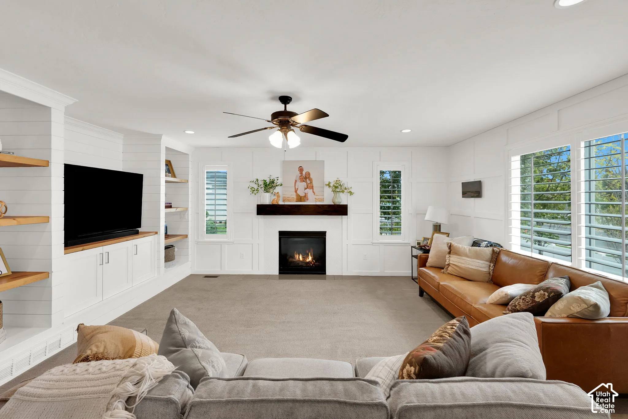 Living room featuring white board-and-batten wall detailing, custom built-in cabinetry with wood shelving, a fireplace with a dark timber mantel, recessed ceiling lighting, and plantation shutters