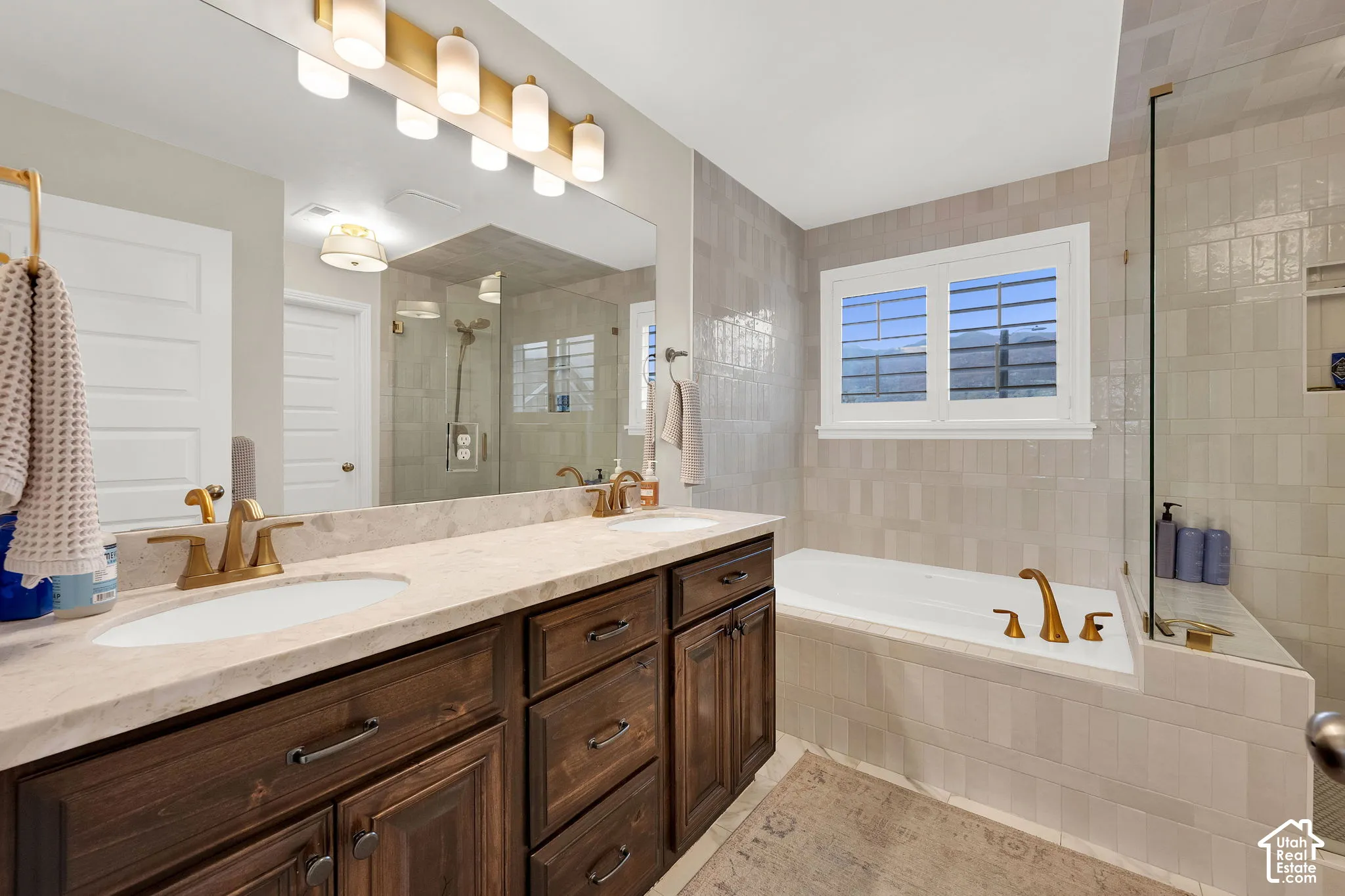 Dual-sink vanity with stone countertop and brass-finish fixtures, inset soaking tub with tile surround, and frameless glass shower enclosure