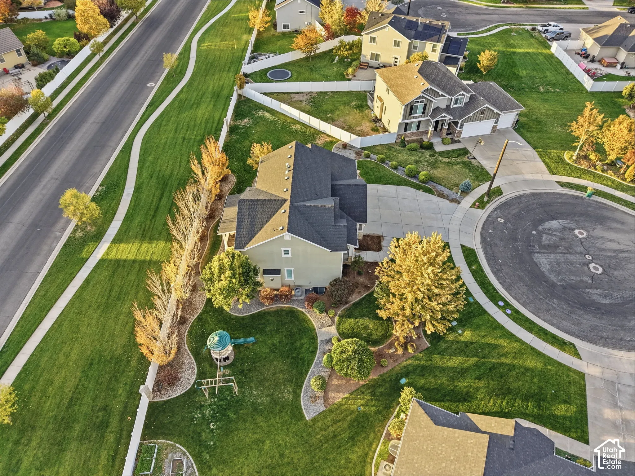 Aerial perspective of a multi-level residence featuring a concrete driveway, asphalt shingle roof, landscaped lawn with curved stone edging, and mature trees