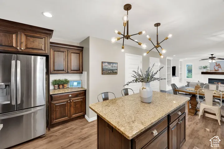 Kitchen featuring dark wood-finish cabinetry, stone countertops, a central island, stainless steel refrigerator, and dual geometric chandeliers