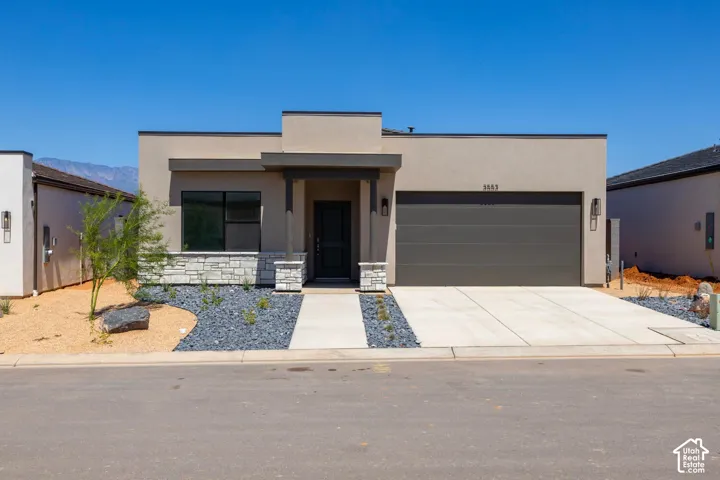 Contemporary single-story exterior featuring smooth stucco siding, stone veneer accents, a flat roofline, a concrete driveway, and mountain scenery