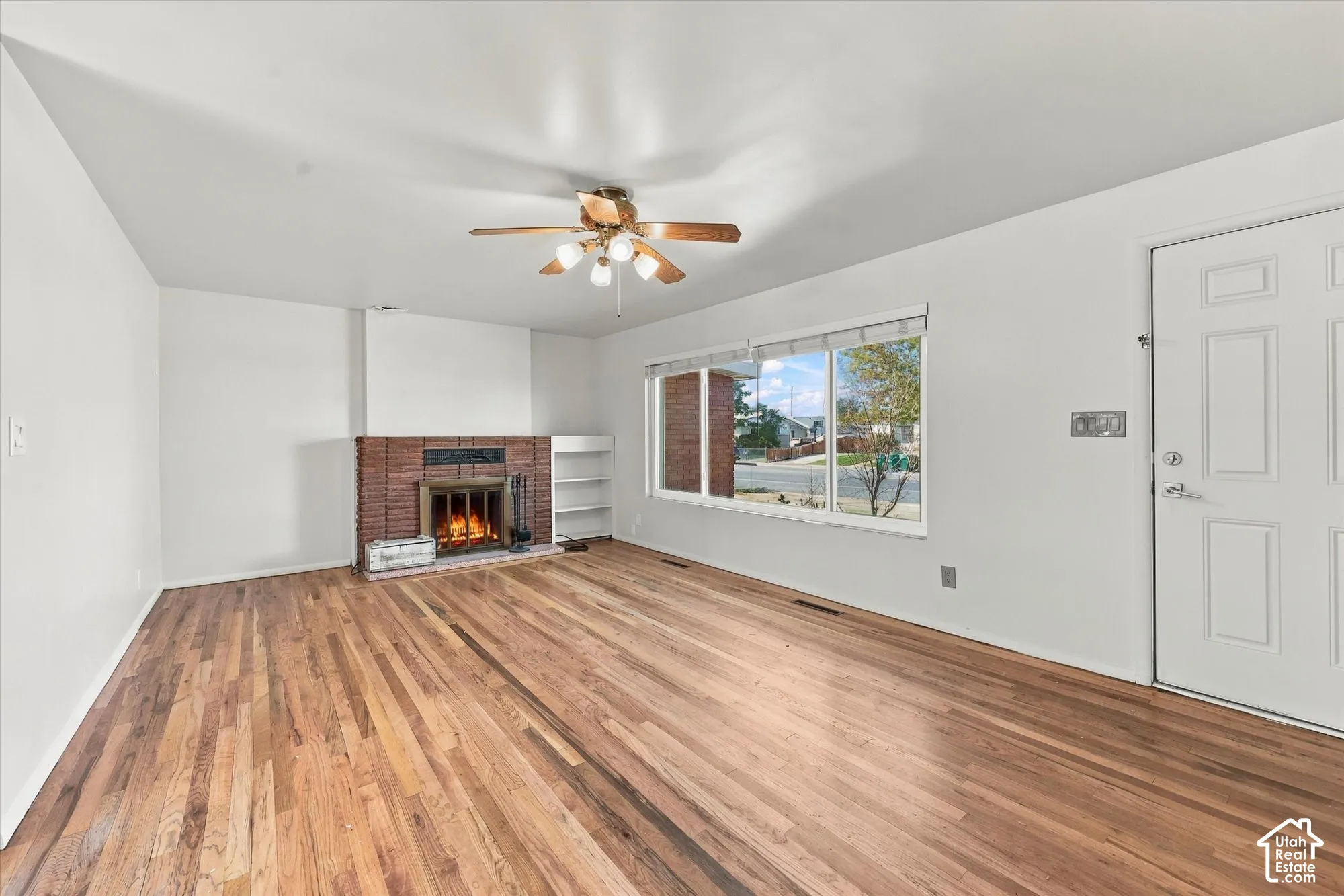 Living area featuring wood-finish flooring, a floor-to-ceiling brick fireplace with a built-in shelf, a large picture window, and a ceiling fan