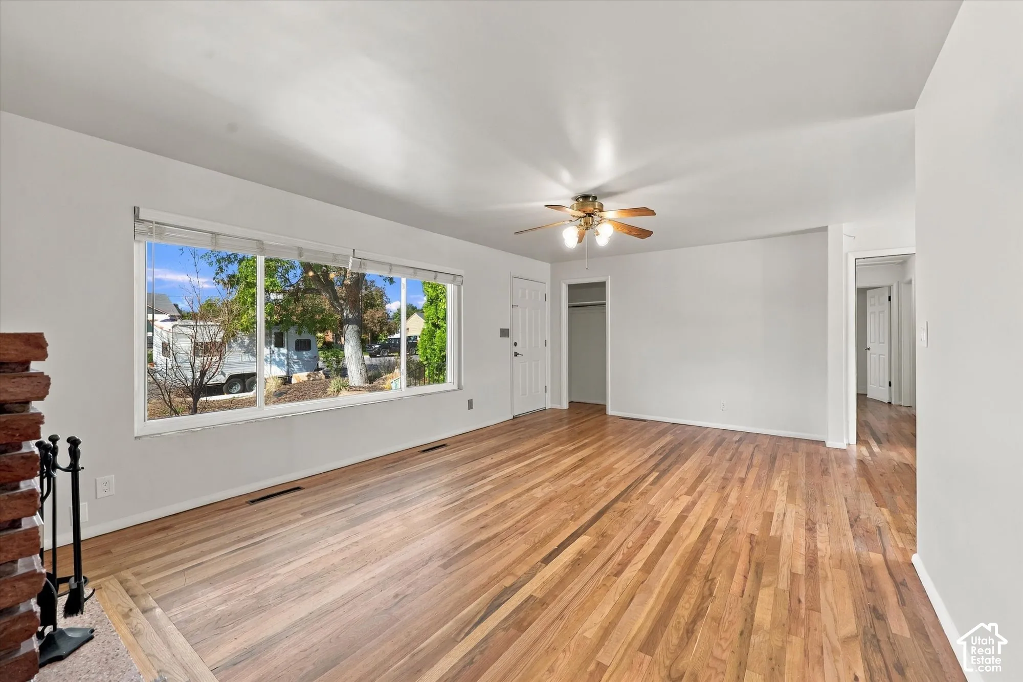 Living area featuring wood-finish flooring, a floor-to-ceiling brick fireplace surround, a large picture window, and a ceiling fan with integrated lighting