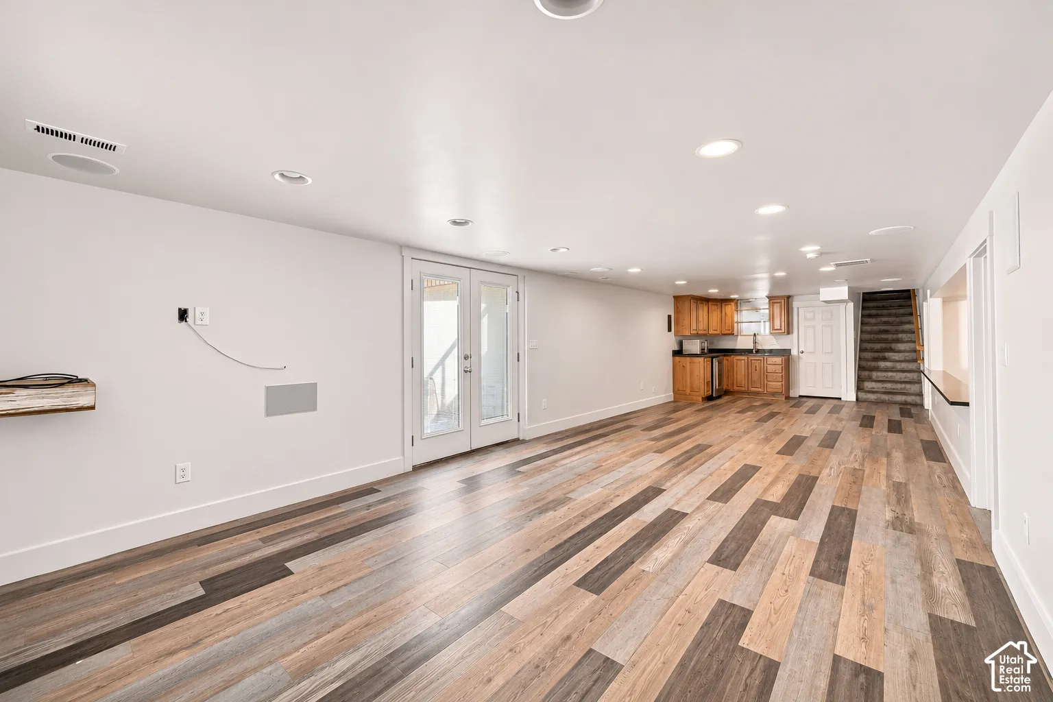 Multipurpose area featuring wood-finish flooring, recessed lighting, double glass-panel doors, kitchenette with cabinetry, and an interior staircase