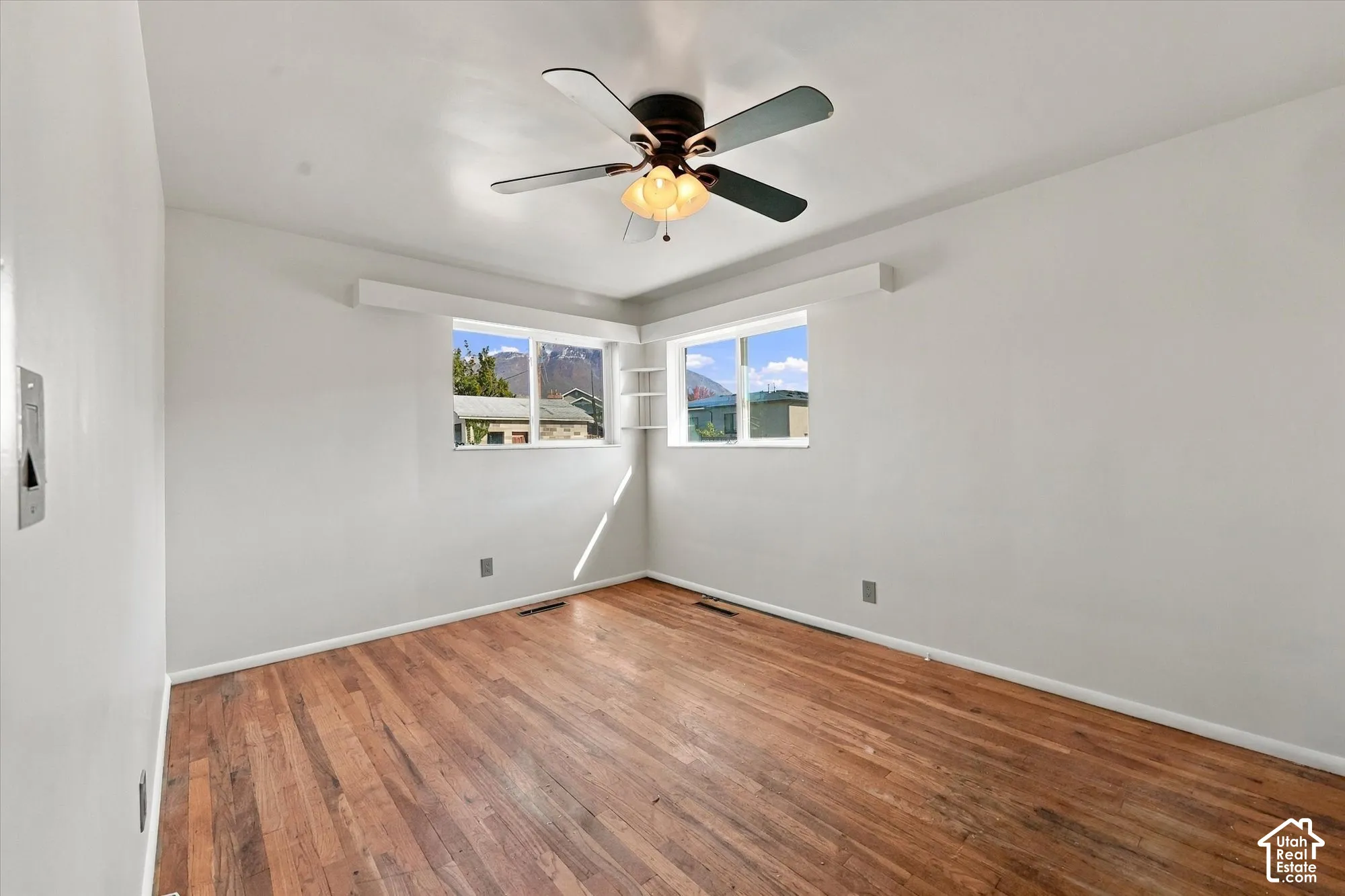 Room featuring wood-finish flooring, dual-aspect windows, a multi-blade ceiling fan with integrated lighting, and built-in corner shelving