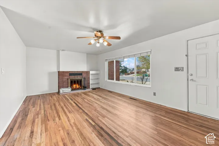 Living area featuring wood-finish flooring, a floor-to-ceiling brick fireplace with a built-in shelf, a large picture window, and a ceiling fan