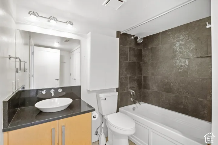 Bathroom featuring a vanity with a dark countertop and vessel sink, large wall-mounted mirror, light wood-finish cabinetry, and a bathtub with full-height dark tiled surround