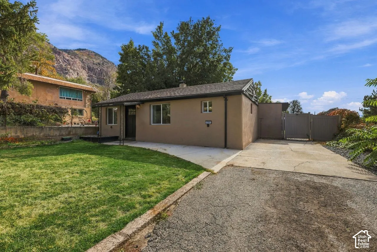 Single-story exterior with stucco siding, composition shingle roof, concrete driveway, attached gate, and mountain backdrop