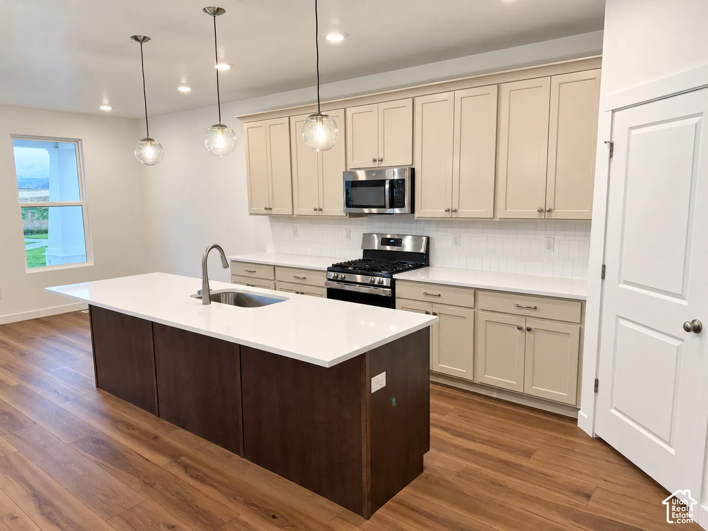 Kitchen featuring a central island with a solid-surface countertop and undermount sink, shaker-style cabinetry, stainless steel appliances, white tile backsplash, and wood-finish flooring
