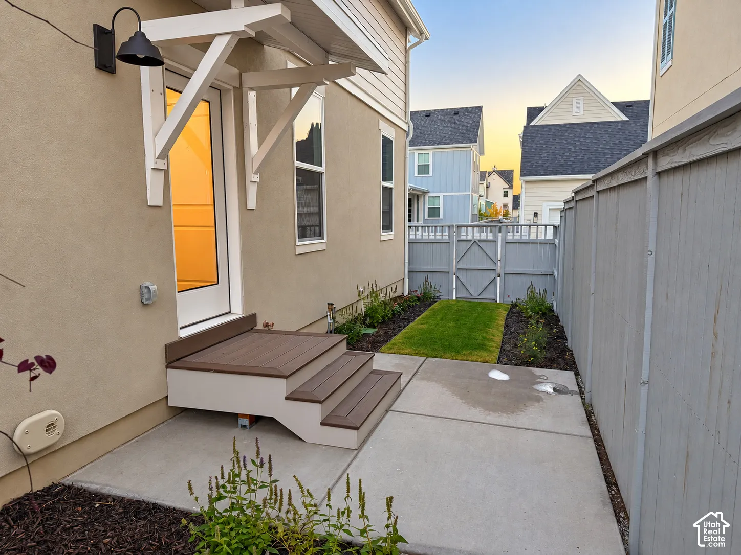 Exterior side patio featuring a concrete slab, composite steps with an overhead bracketed canopy, fenced perimeter, and landscaped beds with integrated turf