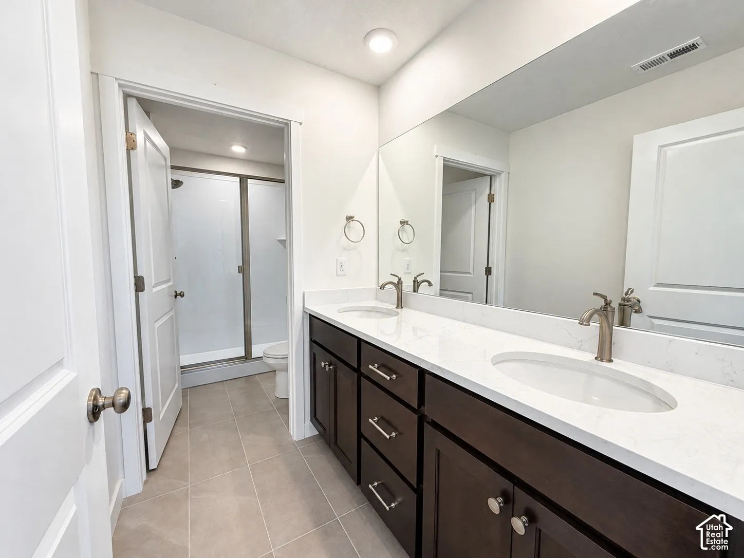 Dual-sink vanity with stone-finish countertop, dark-toned cabinetry, full-width wall mirror, and porcelain tile flooring