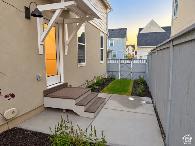 Exterior side patio featuring a concrete slab, composite steps with an overhead bracketed canopy, fenced perimeter, and landscaped beds with integrated turf