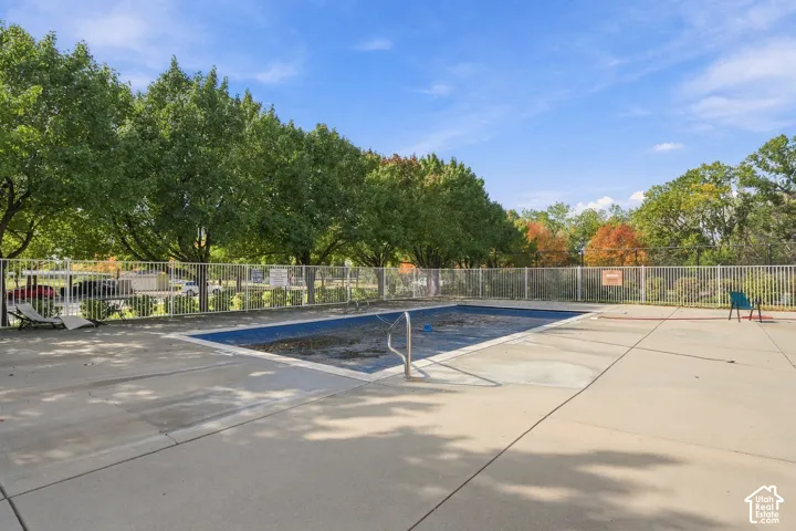 Rectangular inground pool with metal handrail and concrete surround, enclosed by a white metal perimeter fence and mature trees