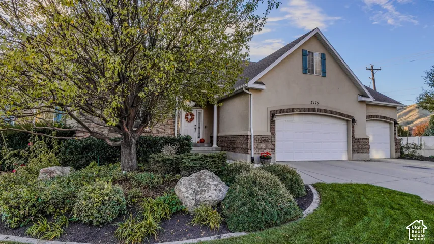 Stucco and stone veneer exterior featuring a three-car garage, covered entryway, and landscaped front yard with mature trees