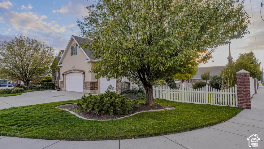 Exterior facade featuring stone veneer siding, multi-car garage, manicured lawn with mature landscaping, and a white picket fence anchored by brick masonry pillars