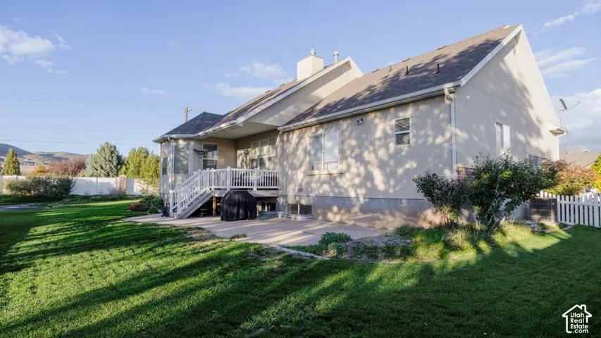 Exterior featuring a stucco facade, elevated wood deck, concrete patio, white vinyl fencing, and grass lawn