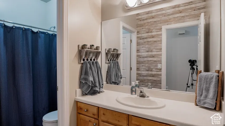 Bathroom featuring a wood-finish vanity with a molded countertop, integrated oval basin, full-width vanity mirror, and accent wall with horizontal plank-style finish