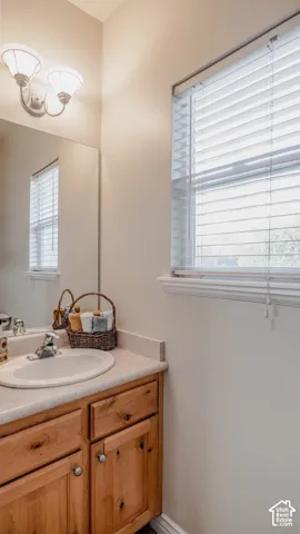 Vanity featuring wood cabinetry, a light-toned countertop, an integrated oval sink, and a dual-light wall sconce, complemented by a large wall-mounted mirror and a window with horizontal blinds