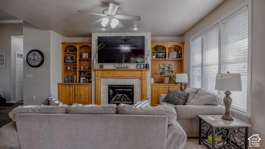 Fireplace with a wood mantle and tile surround, flanked by custom built-in cabinetry and shelving, ceiling fan, and large windows with horizontal blinds