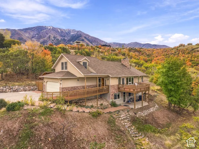 Multi-level residence featuring stone and stucco exterior, attached two-car garage, elevated wood deck, stone masonry chimney, and stone hardscape paths set against a mountain backdrop