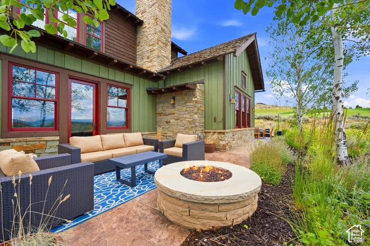 Exterior patio featuring a circular stone fire pit, stamped concrete flooring, board-and-batten siding, stone chimney masonry, and double-hung windows with red trim