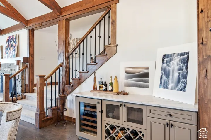 Built-in cabinetry featuring a beverage refrigerator, wine rack, and light-toned countertop, complemented by exposed timber beams and an open staircase with decorative metal balusters