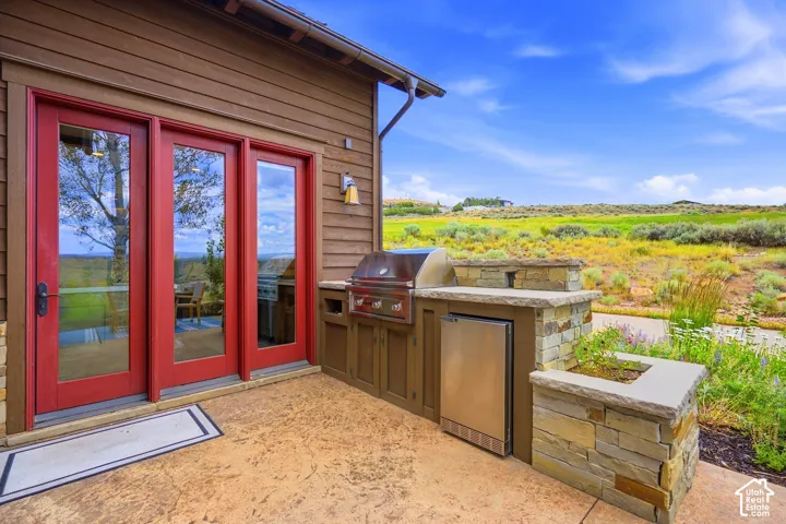 Covered patio featuring stamped concrete flooring, a built-in outdoor kitchen with a stainless steel grill, integrated refrigeration, stone masonry, and red-trimmed glass paneled doors