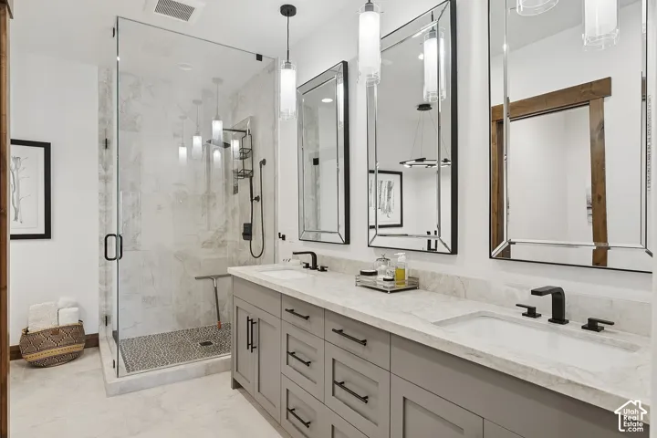 Dual-vanity cabinetry with stone countertop and matte black fixtures, walk-in glass-enclosed shower featuring tiled surround and pebble-texture flooring, and decorative wall mirrors with metallic frames