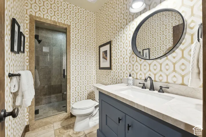Custom blue vanity with light-toned stone countertop, integrated undermount sink, patterned wall covering, oval wall-mounted mirror, and glass-enclosed shower with stone tile surround