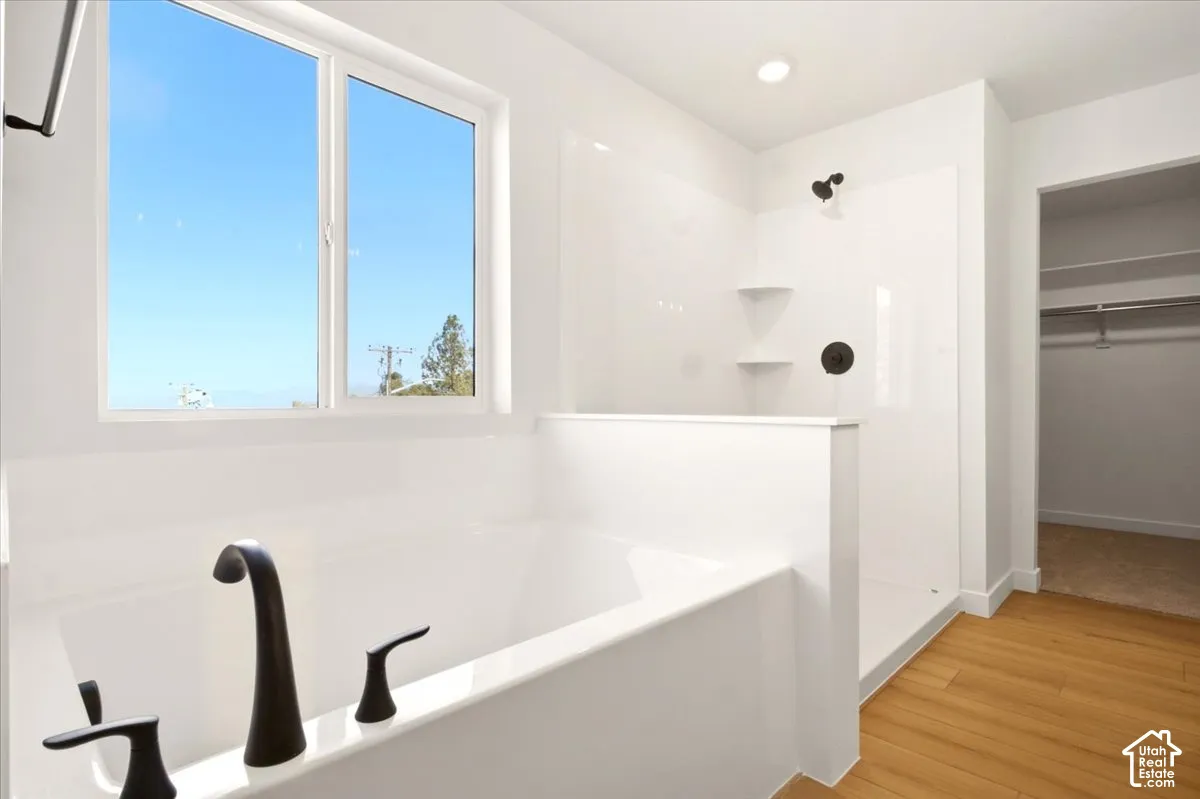 Deep soaking tub and separate shower featuring dark hardware, large window, and wood-finish flooring